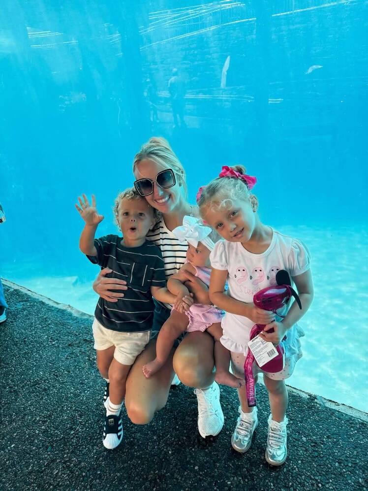 Brittany Mahomes poses in front of a large aquarium window while holding baby Golden and crouching beside her older children, Bronze and Sterling. Bronze waves at the camera in a striped shirt and white shorts, while Sterling holds a toy fan and wears a white outfit with a red bow in her hair.