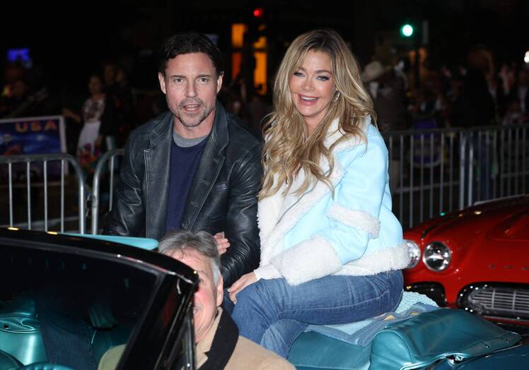 Denise Richards and Aaron Phypers ride together in the back of a convertible at a nighttime event. Richards is smiling in a light blue shearling jacket and jeans, while Phypers wears a black leather jacket over a dark shirt.