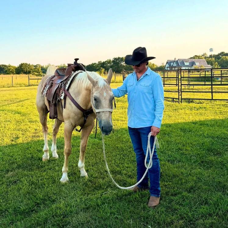 Josh Hall wears a cowboy hat and light blue button-up shirt while standing in a grassy field, holding the reins of a saddled tan horse.