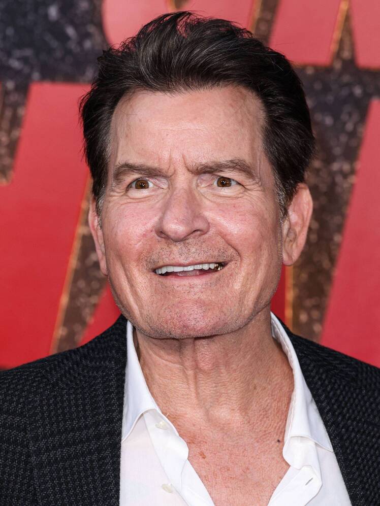 Charlie Sheen smiles on the red carpet, wearing a black textured blazer over a white shirt.